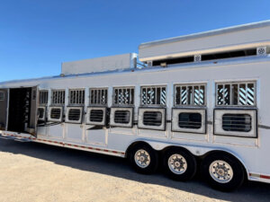 A large white livestock trailer with multiple barred windows and three axles is parked on gravel under a clear blue sky. The back door is open, revealing the interior. -Stillwater Trailer Sales