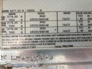 A close-up of a vehicle information sticker showing details like GVWR, GAWR, tire size (LT235/85R16E), rim size (16x7J), PSI, and conformity statements in English and French. The VIN is partially visible at the bottom. -Stillwater Trailer Sales