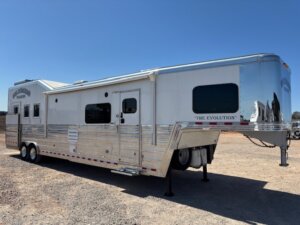 A large, white fifth-wheel trailer labeled The Evolution is parked on gravel under a clear blue sky. The trailer has multiple windows, silver detailing, and multiple wheels for support. -Stillwater Trailer Sales