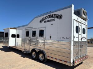 A large silver Bloomer horse trailer with multiple windows and a slide-out section is parked on gravel under a clear blue sky. -Stillwater Trailer Sales