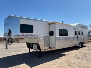 A large, silver gooseneck horse trailer with multiple windows and &ldquo;The Evolution&rdquo; written on the side is parked on gravel under a clear blue sky. -Stillwater Trailer Sales