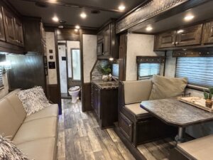 Interior of an RV featuring a beige sofa, a dining area with a marble-look table, dark wood cabinets, wood-look flooring, and a view of a bathroom with a toilet in the background. -Stillwater Trailer Sales