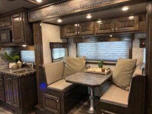 Cozy RV interior with a kitchenette on the left and a dining nook on the right, featuring a marble-patterned table, cushioned bench seating, throw pillows, and potted plants for decor. Warm lighting highlights dark wood cabinets. -Stillwater Trailer Sales