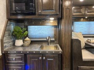 A compact kitchen area with dark wood cabinets, a small sink, a microwave, potted plants, and decorative jars. A dining nook with cushioned seats is visible to the right under a window with closed blinds. -Stillwater Trailer Sales