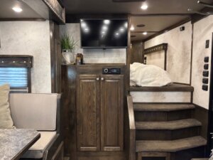 Cozy RV interior with wooden cabinets, a TV mounted above a stereo, stairs leading to a loft bed with a white blanket, a plant on the shelf, and a dining area with cushioned seating on the left. -Stillwater Trailer Sales