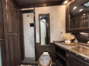 A bathroom with dark wood cabinets, a textured wall, a narrow vertical mirror with metal studs, a toilet, a countertop with a gold sink, and a decorative vase with dried plants. Warm lighting illuminates the space. -Stillwater Trailer Sales