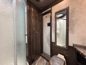 A small bathroom with a glass shower door, wooden cabinets, a toilet with a patterned lid, and a tall mirror with metal studs along the edge. The walls have a textured, light-colored finish. -Stillwater Trailer Sales