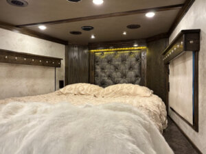 Luxurious RV bedroom with a large, plush bed covered in cream bedding and a faux fur throw. The room features a tufted headboard, wood accents, built-in lights, and a cozy, upscale atmosphere. -Stillwater Trailer Sales