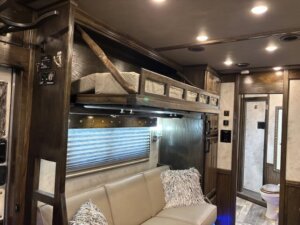 A modern RV interior with a beige sofa, decorative pillows, a fold-down bunk bed above, wood paneling, and a view into a bathroom with a toilet and mirror in the background. Warm ceiling lights illuminate the space. -Stillwater Trailer Sales