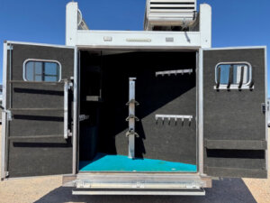 The open back doors of a horse trailer reveal a clean, carpeted interior with black walls, multiple hooks for tack, and saddle racks in the center, all under a clear blue sky. -Stillwater Trailer Sales