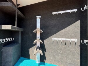 Inside a storage area with grey carpeted walls, multiple rows of metal hooks, and a vertical rack holding three black saddles. The floor is covered with bright blue carpet. -Stillwater Trailer Sales