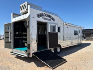 A large white Bloomer horse trailer with an open rear ramp and side door, revealing stalls and storage compartments inside, parked on gravel under a clear blue sky. -Stillwater Trailer Sales