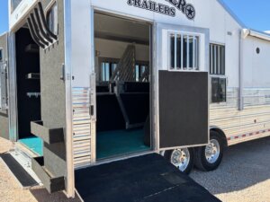 A silver horse trailer with open doors shows padded stalls inside, non-slip flooring, and a black ramp extending to the ground for easy animal entry. Sunlight highlights the trailer&rsquo;s clean, organized interior. -Stillwater Trailer Sales
