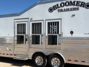 A silver Bloomer horse trailer with three barred windows and two doors, parked under a clear blue sky. The trailer has shiny metallic paneling and two wheels visible. -Stillwater Trailer Sales