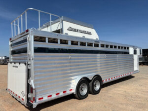 A large silver Elite 8 Horse Trailer with dual axles is parked on gravel under a clear blue sky. The trailer features horizontal slats, a rear loading door, and a roof rack with a white Elite Trailers storage box. -Stillwater Trailer Sales
