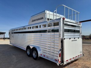 A large silver Elite 8 Horse Trailer with two axles, side vents, a rear loading door, and an upper storage rack is parked on gravel under a clear blue sky. -Stillwater Trailer Sales