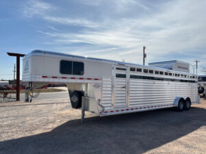 A large silver Elite 8 Horse Trailer with side vents is parked on gravel under a partly cloudy sky, with power lines and a few structures visible in the background. -Stillwater Trailer Sales