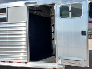Open door of a silver Elite 8 Horse Trailer, revealing a dark, carpeted interior with hooks inside, parked on a gravel surface. -Stillwater Trailer Sales
