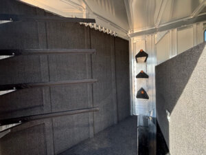 Interior of an Elite 8 Horse Trailer featuring three saddle racks on the left wall, three small bridle hooks on the right, and carpeted flooring complemented by metallic paneling. -Stillwater Trailer Sales