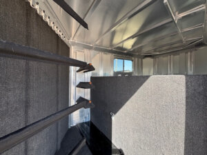 Interior of an Elite 8 Horse Trailer with carpeted walls and dividers, four angled saddle racks on the left, and a small window letting in daylight. The space appears clean and organized. -Stillwater Trailer Sales