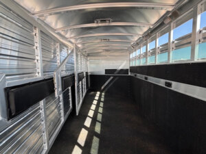 Interior of an empty Elite 8 Horse Trailer with ribbed aluminum walls, black padding along the sides, and sunlight streaming in through open slats on the right. The dark, textured floor completes this spacious setting. -Stillwater Trailer Sales