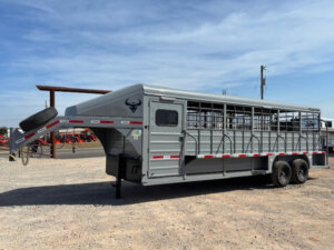 A large gray livestock trailer with open slats and a gooseneck hitch is parked on gravel. It has two axles, a spare tire mounted on the front, and red-and-white reflective strips along the sides. -Stillwater Trailer Sales
