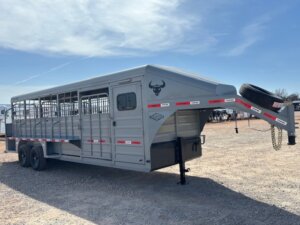 A large gray livestock trailer with a gooseneck hitch, side vents, and a spare tire mounted in front, parked on a gravel lot under a partly cloudy sky. -Stillwater Trailer Sales