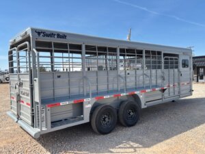 A silver Swift Built livestock trailer with dual axles and side ventilation openings is parked on gravel under a clear sky. -Stillwater Trailer Sales