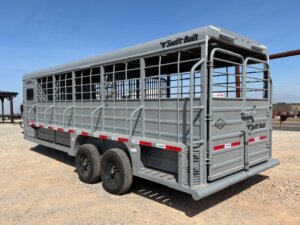 A large gray livestock trailer with barred sides and double axles is parked on gravel. The trailer has Swift Built branding and is designed for transporting animals. Red and white reflective tape runs along the sides. -Stillwater Trailer Sales