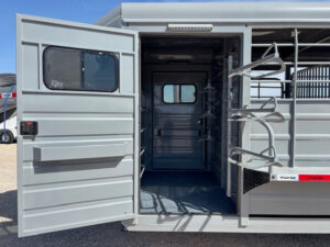 A metal trailer with an open door reveals multiple vertical racks inside, likely for storing saddles or equipment. The interior is gray and well-lit, with a window on the opposite wall. -Stillwater Trailer Sales