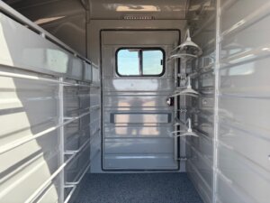 Interior of an empty, clean horse trailer with metal walls, windows on the door, saddle racks attached on the right, and textured flooring. Sunlight shines through the rear window. -Stillwater Trailer Sales