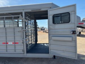 A gray livestock trailer with its side door open, revealing the interior and divider gates. The trailer is parked on gravel with other trailers visible in the background under a clear blue sky. -Stillwater Trailer Sales