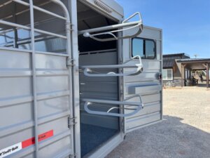 The image shows the open back door of a silver horse trailer with three empty saddle racks mounted on the inside of the door. A building and blue sky are visible in the background. -Stillwater Trailer Sales