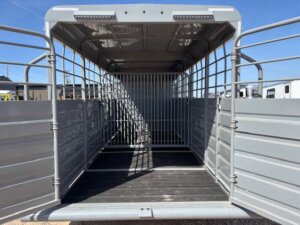 Open, empty livestock trailer with metal sides and roof, black ribbed flooring, and a gated divider in the center. The rear doors are open, showing the trailer&rsquo;s spacious and well-lit interior under a blue sky. -Stillwater Trailer Sales