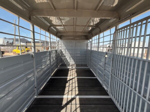 Interior view of an empty livestock trailer with metal walls, slatted floor, and grated gates at the far end. Sunlight casts shadows through the grates. Industrial buildings and equipment are visible outside. -Stillwater Trailer Sales