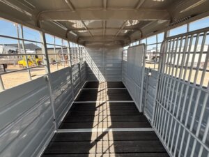 Interior view of an empty livestock trailer with metal walls, grated gates on both sides, and a textured floor, designed for safely transporting animals. Sunlight shines in from openings along the upper sides. -Stillwater Trailer Sales