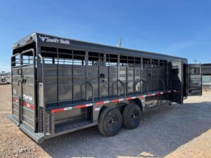 A black Swift Built livestock trailer with two axles and side rails is parked on gravel under a clear blue sky. The rear and side gates are open, showing the interior compartments. -Stillwater Trailer Sales