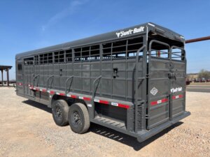 A large, black Swift Built livestock trailer with two axles is parked on a gravel lot under a clear blue sky. The trailer has open slats, safety reflectors, and branding on the side and back. -Stillwater Trailer Sales