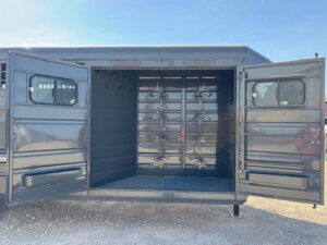 Open black metal trailer with two wide doors, showing an empty interior and multiple vertical racks inside, parked on gravel under a bright sky. -Stillwater Trailer Sales
