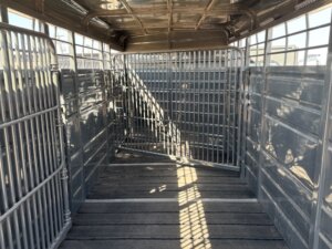 Interior of an empty metal livestock trailer with grated gates and wooden slatted floor, showing bars and sunlight casting shadows inside the trailer. -Stillwater Trailer Sales