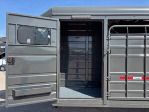 A gray livestock trailer with its side door open, revealing a carpeted step and an empty interior. The trailer is parked outdoors on a sunny day. -Stillwater Trailer Sales