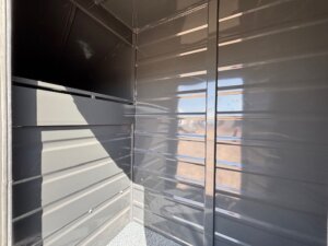 Interior view of a metal structure with smooth, reflective gray walls and a corner where sunlight streams in, casting shadows and a partial reflection of an outdoor scene on the right wall. -Stillwater Trailer Sales