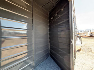 Interior of an empty metal trailer with ribbed dark walls, a small pebbled floor area, and an open door revealing a sunny outdoor lot with other trailers in the background. -Stillwater Trailer Sales