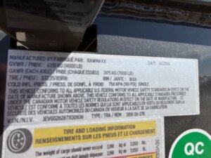 Close-up of a vehicle information sticker displaying manufacturer details, weight limits, tire size (ST215/75D15), rim size (15x6), cold tire pressure (110 PSI), and a vehicle identification number (VIN). -Stillwater Trailer Sales