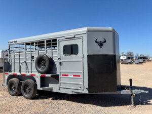 A gray livestock trailer with a black bull head logo is parked on gravel under a clear blue sky. The trailer has two axles, a spare tire mounted on the side, and a small window on the front. -Stillwater Trailer Sales