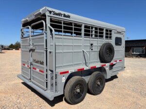 Gray Swift Built livestock trailer with barred sides, two axles, a spare tire mounted on the side, and red-and-white reflective tape, parked on a gravel lot under a clear blue sky. -Stillwater Trailer Sales