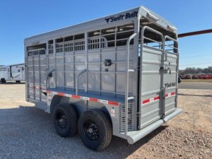 A silver Swift Built livestock trailer with double axles, black wheels, and metal bars, parked on gravel near a road on a clear day. -Stillwater Trailer Sales