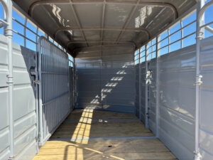 Interior view of an empty metal livestock trailer with wooden flooring and open roof slats, allowing sunlight to create shadows inside. The trailer walls and gate are grey, and the sky is visible through the top. -Stillwater Trailer Sales