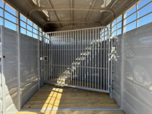 Interior of an empty livestock trailer with metal walls, a wooden floor, and a metal gate separating the front and back sections. Sunlight casts shadows through the open top panels. -Stillwater Trailer Sales