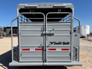 Rear view of a gray Swift Built livestock trailer with double doors, metal bars at the top, red and white reflective tape, and a bull logo, parked on gravel with other trailers and blue sky in the background. -Stillwater Trailer Sales